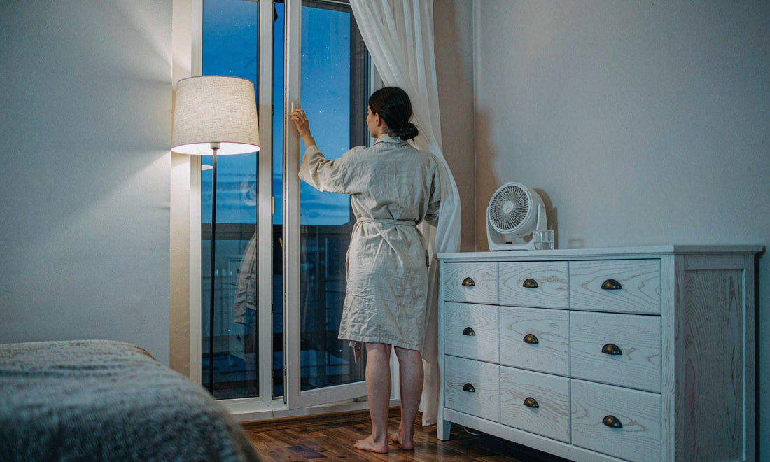 Woman seen from behind opening a tall window at dusk with a fan on the dresser
