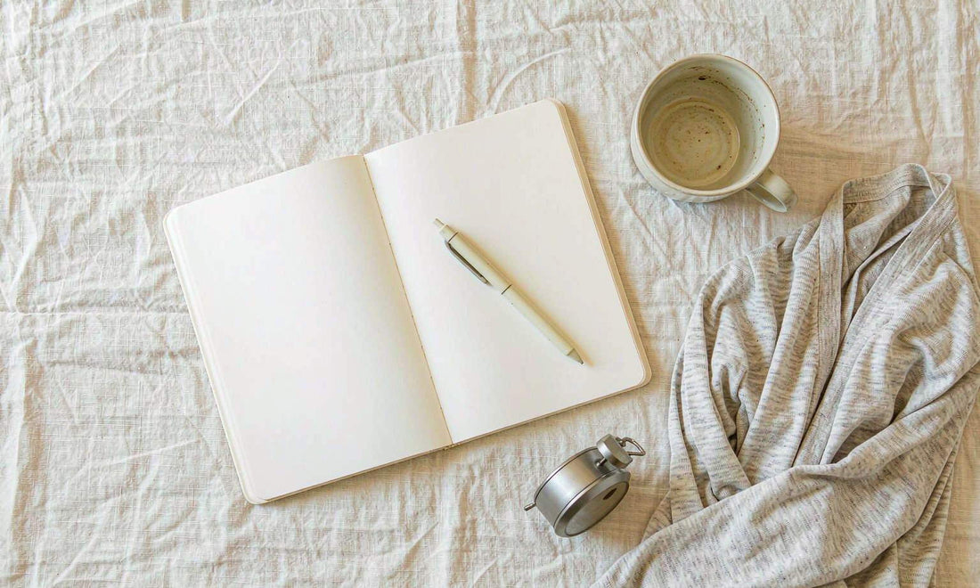 Overhead flat lay of an open notebook, pen, cup and folded cardigan on pale linen bedding