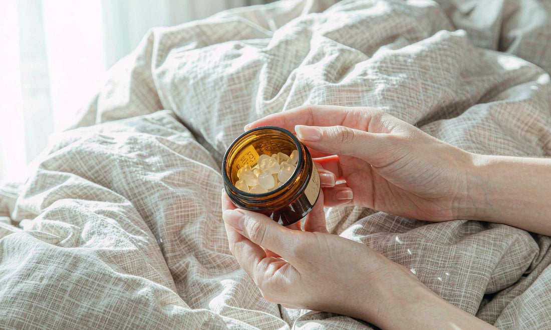 Fair-skinned hands holding a small amber jar of magnesium gummies over a linen duvet.