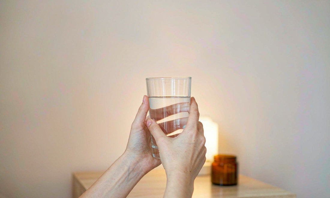 Fair-skinned hands holding a glass of water beside a warm lamp and a small amber jar on a bedside table