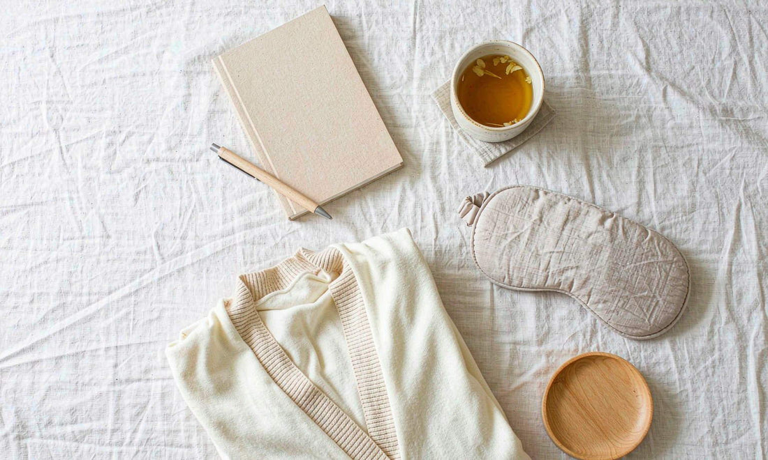 Overhead view of a bedside flat-lay with a notebook, herbal tea, sleep mask and folded cardigan on linen