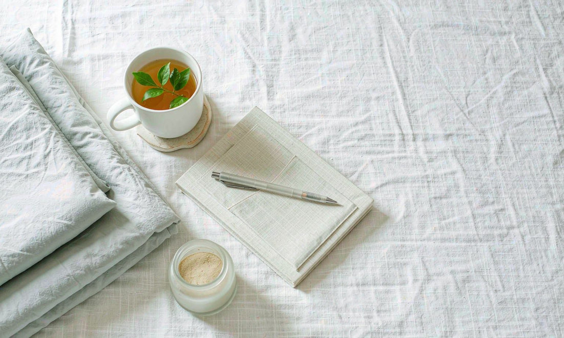 Flat lay of folded bedding, herbal tea, a notebook and a small jar on white linen