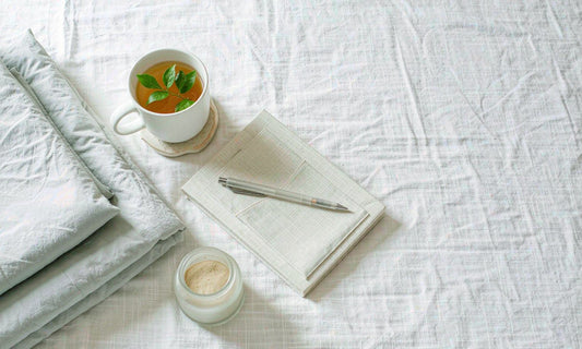 Flat lay of folded bedding, herbal tea, a notebook and a small jar on white linen
