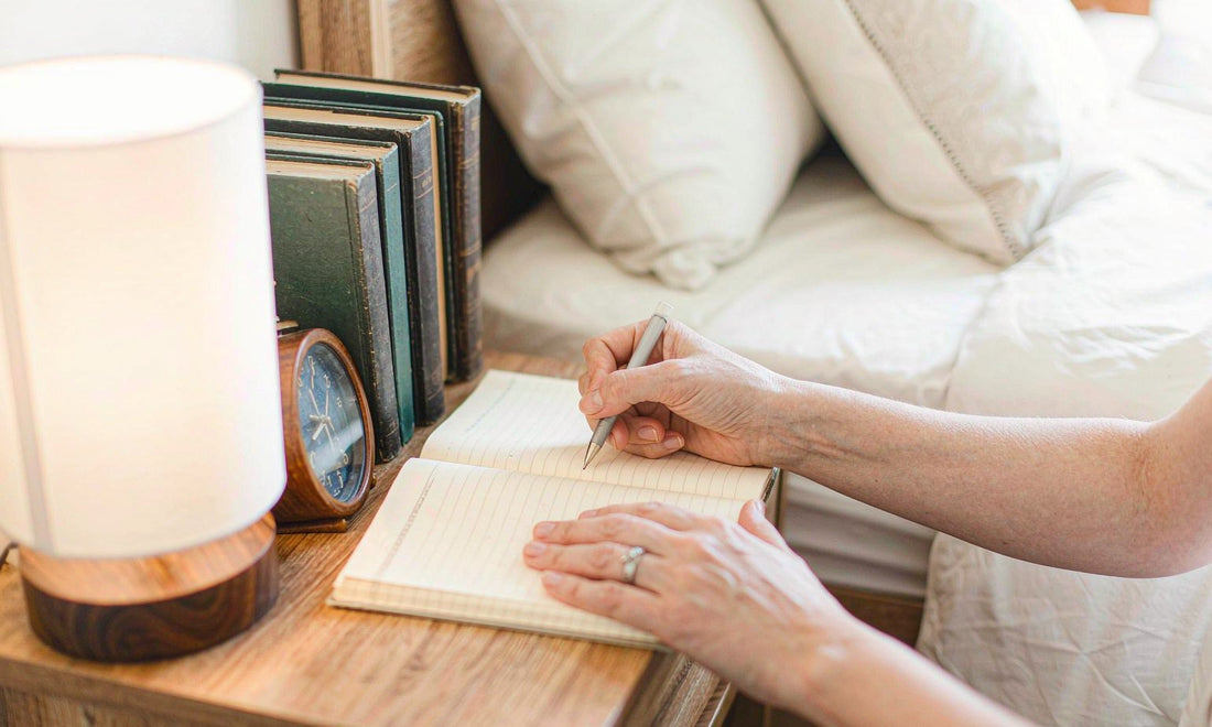 Hands writing in a journal on a nightstand, warm lamp glow