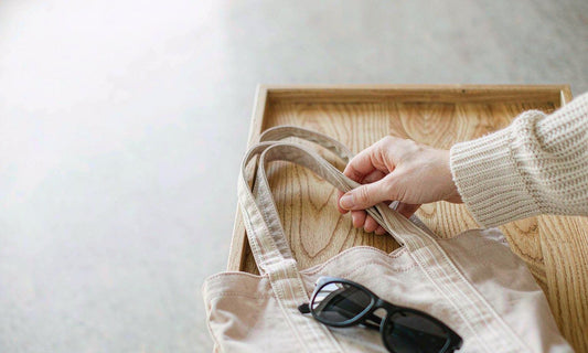 A hand in a cream knit sleeve lifting a canvas tote bag with sunglasses from a light oak bench
