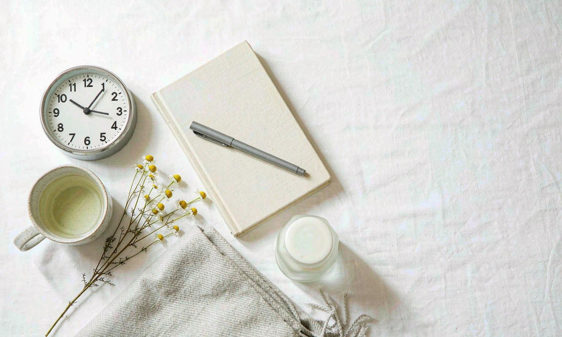Flat lay of a bedside clock, herbal tea, notebook and folded blanket on white linen
